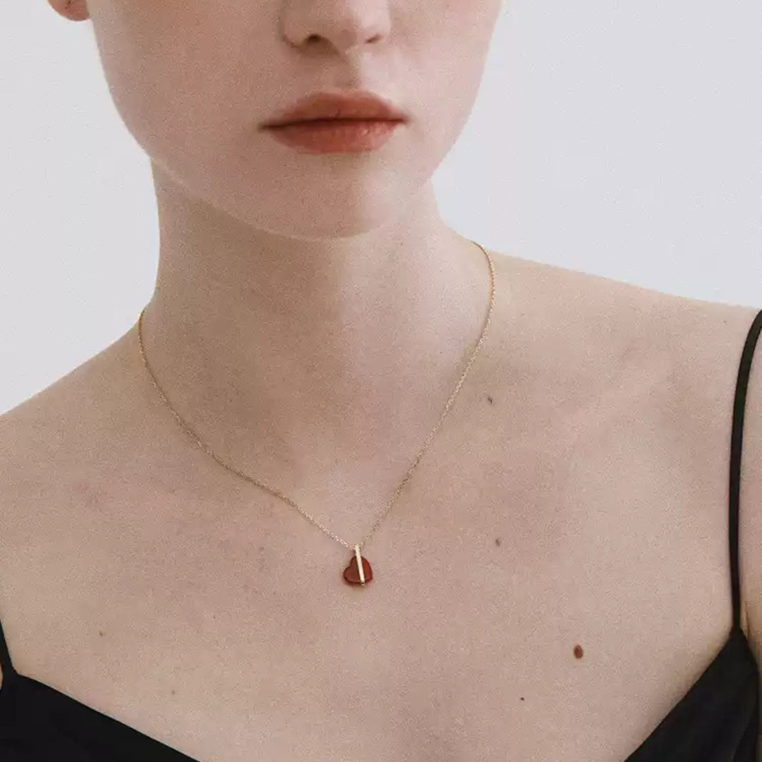 A woman wearing a delicate 14K gold necklace with a red heart pendant on a plain background