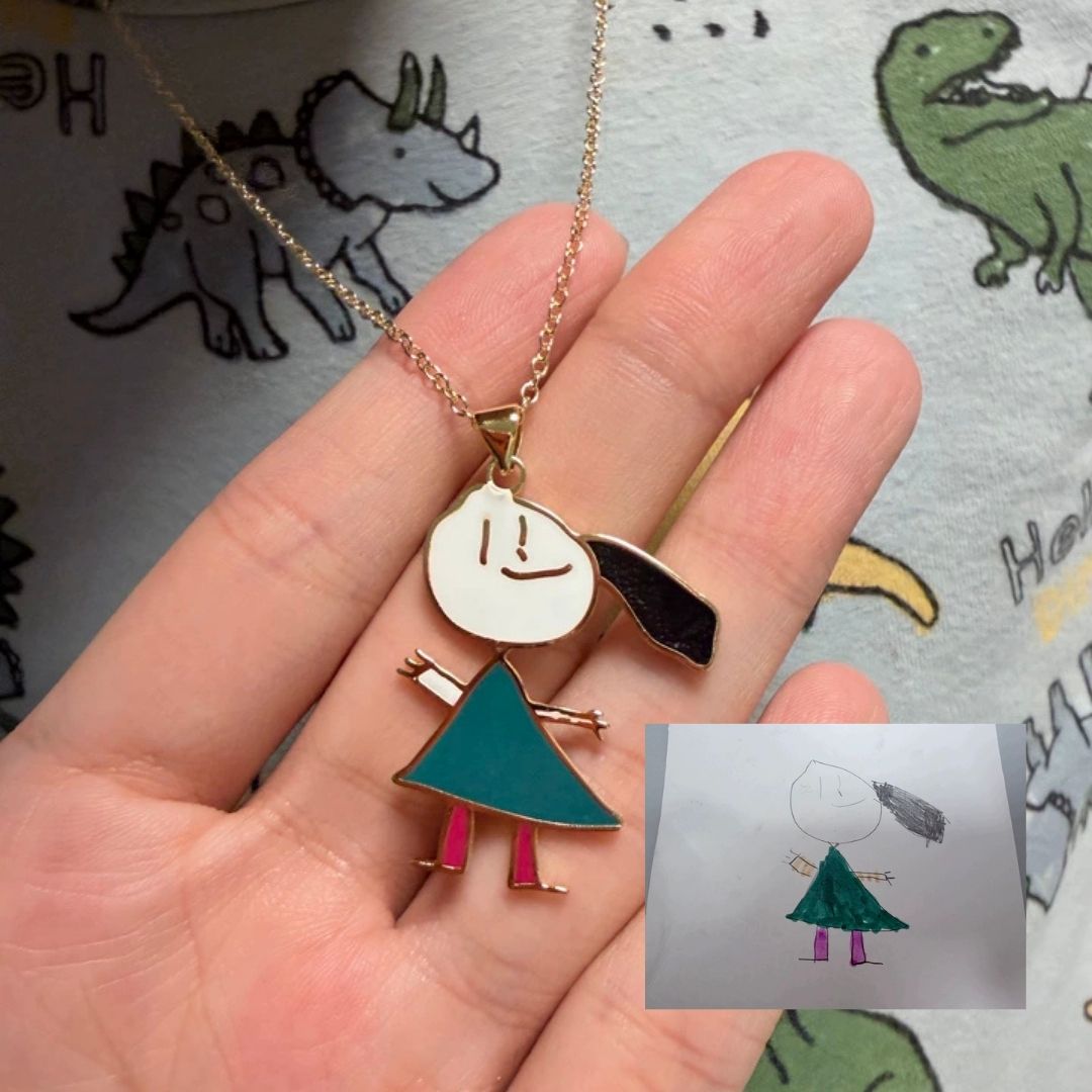 Hand holding a necklace with a pendant shaped like a child, next to a small card with a similar drawing, on a background with dinosaur illustrations.