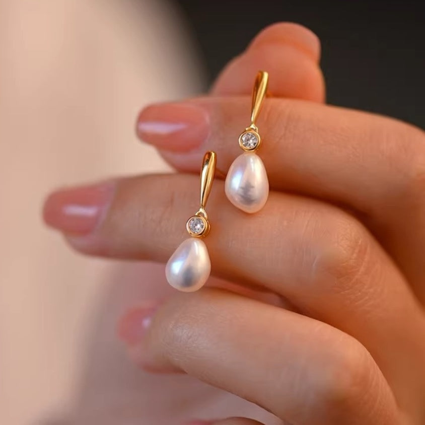 Close-up view of a pair of baroque pearl drop earrings with a geometric gold bar design and zirconia accent held in fingers. 1