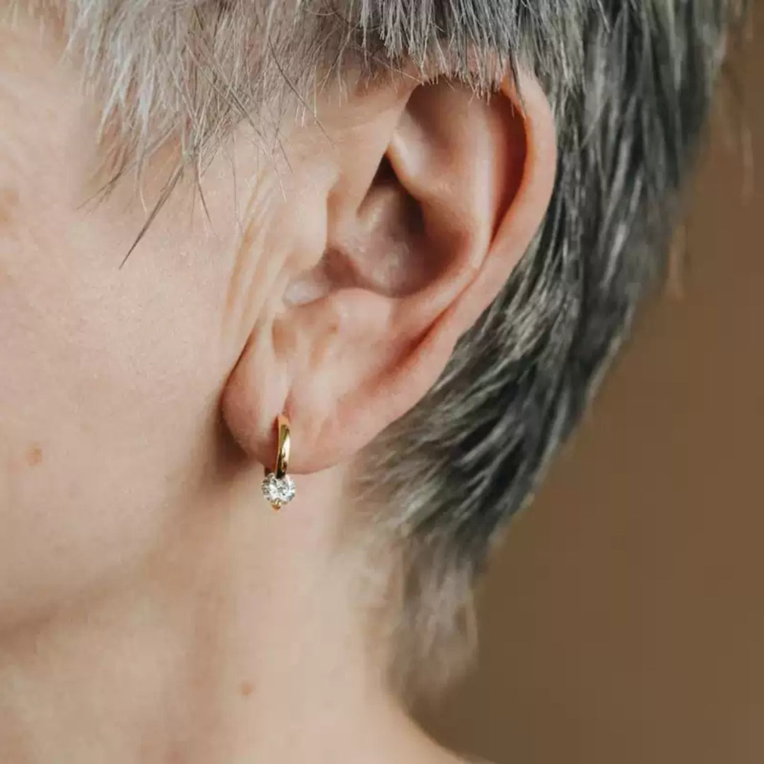 Close-up of an ear wearing a small gold hoop earring with a diamond on a neutral background