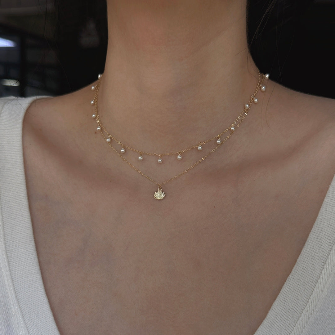 Close-up of a person wearing a delicate gold pearl choker necklace layered with a small gold shell pendant necklace.