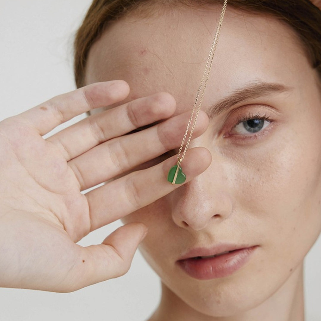 Person holding a gold necklace with a green heart pendant made of jade in front of their face.